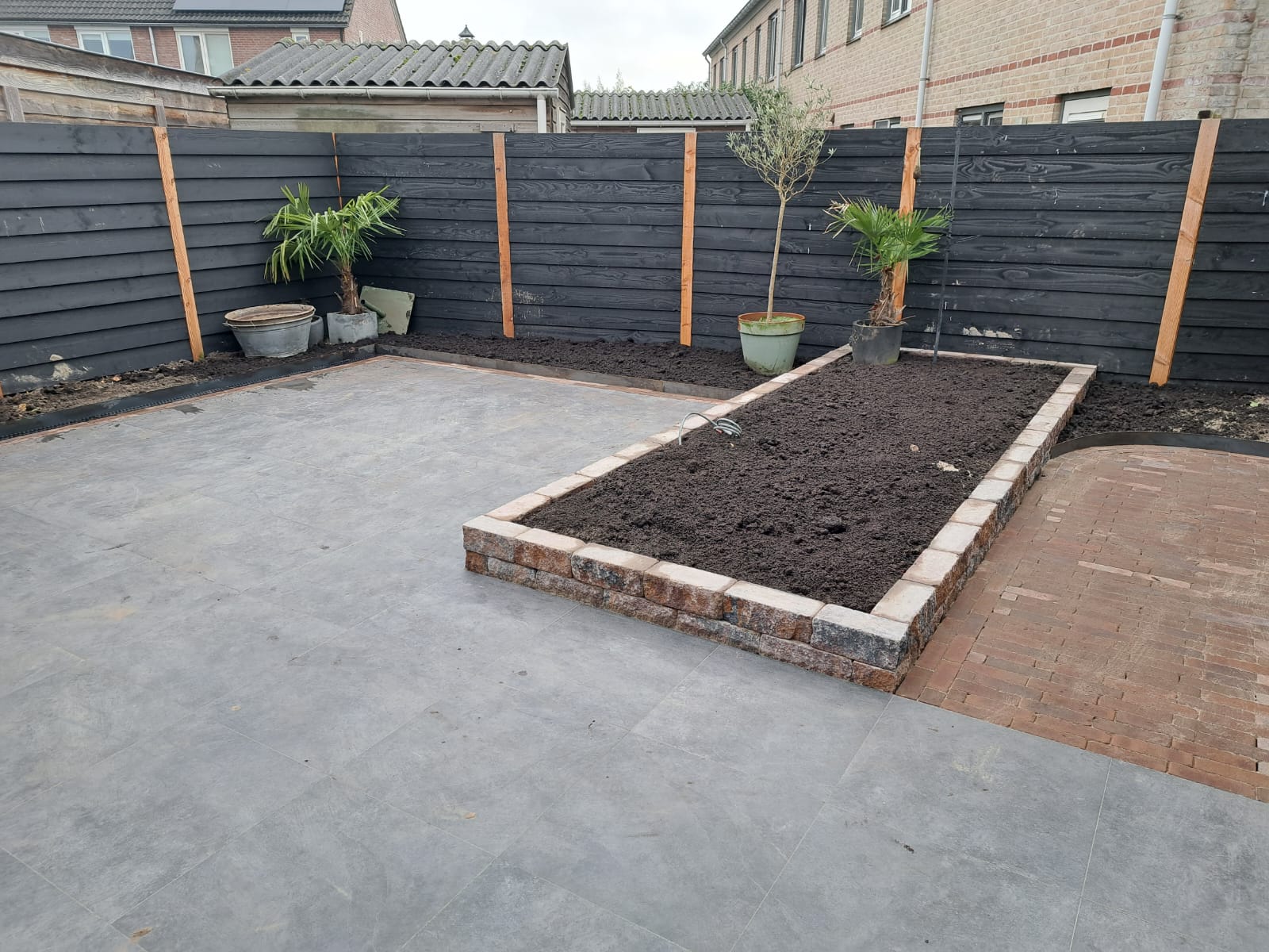 Nieuw terras van grote grijze tegels met een verhoogde border in gestapelde steen en een zwarte schutting