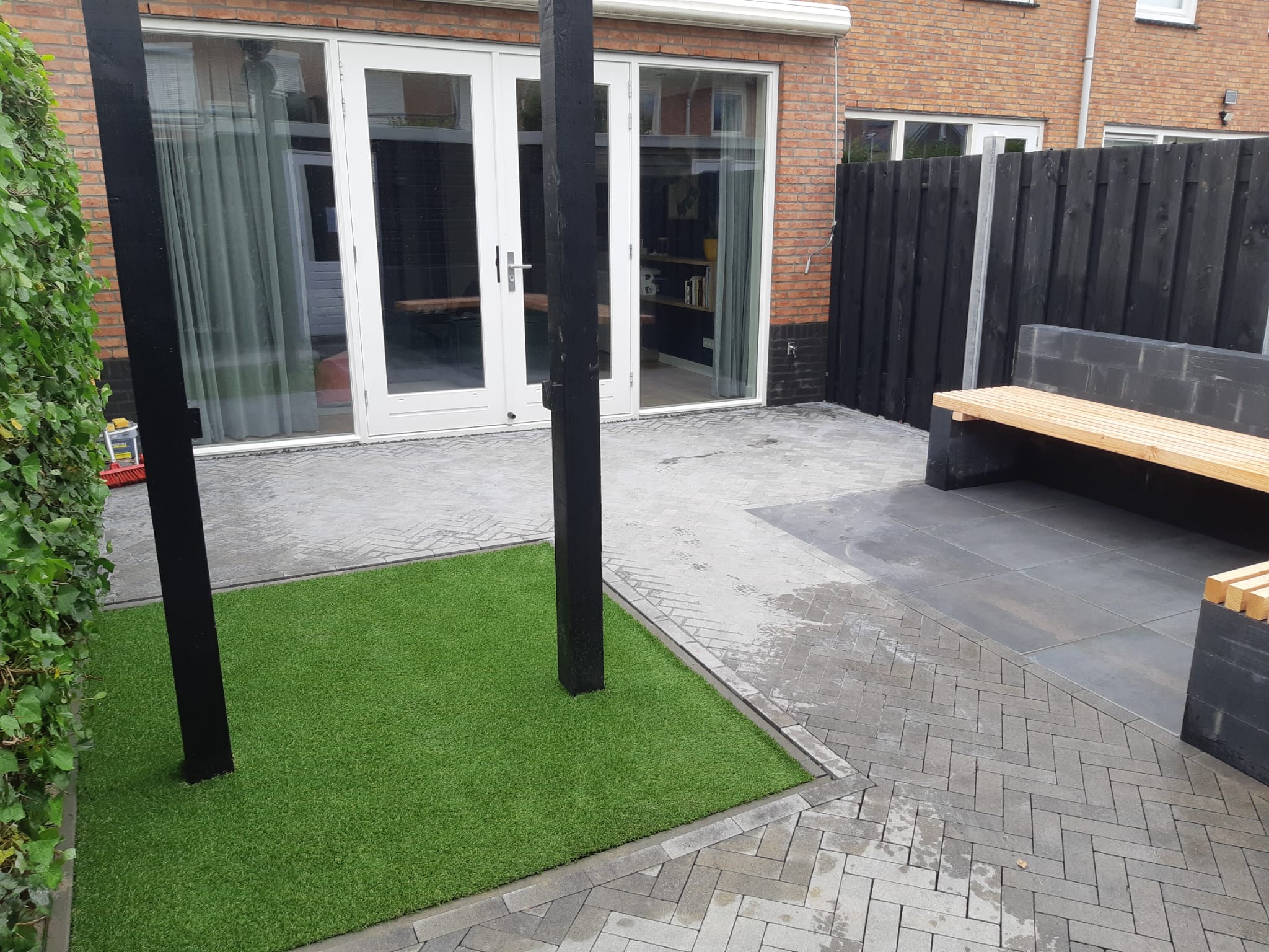 Vak kunstgras tussen visgraat bestrating, zwarte pergolapalen en een zitbank met houten zitting