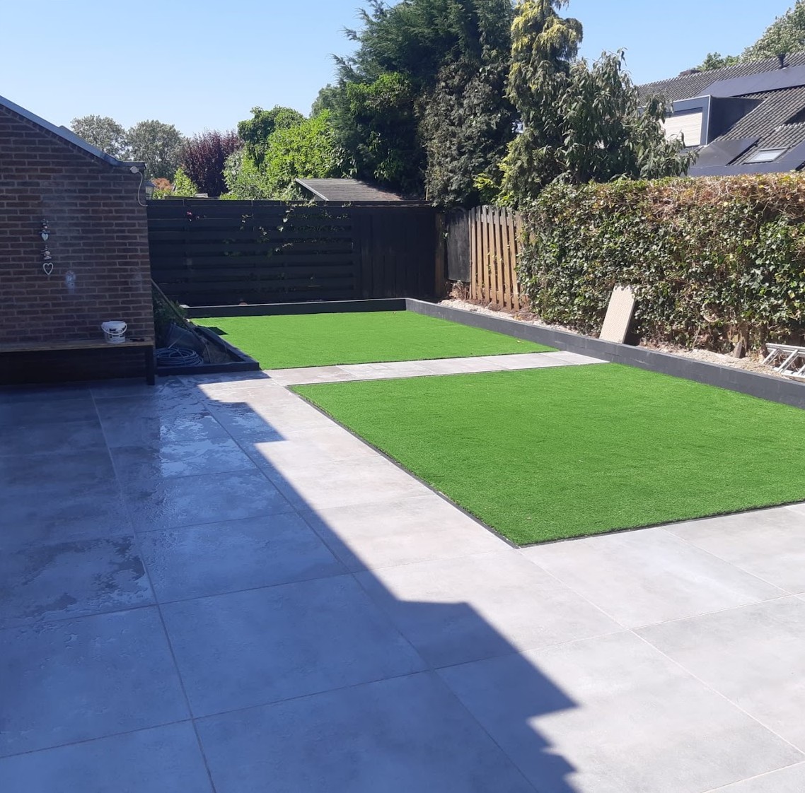Dezelfde tuin na de aanleg: terras van grote grijze tegels met twee vakken kunstgras