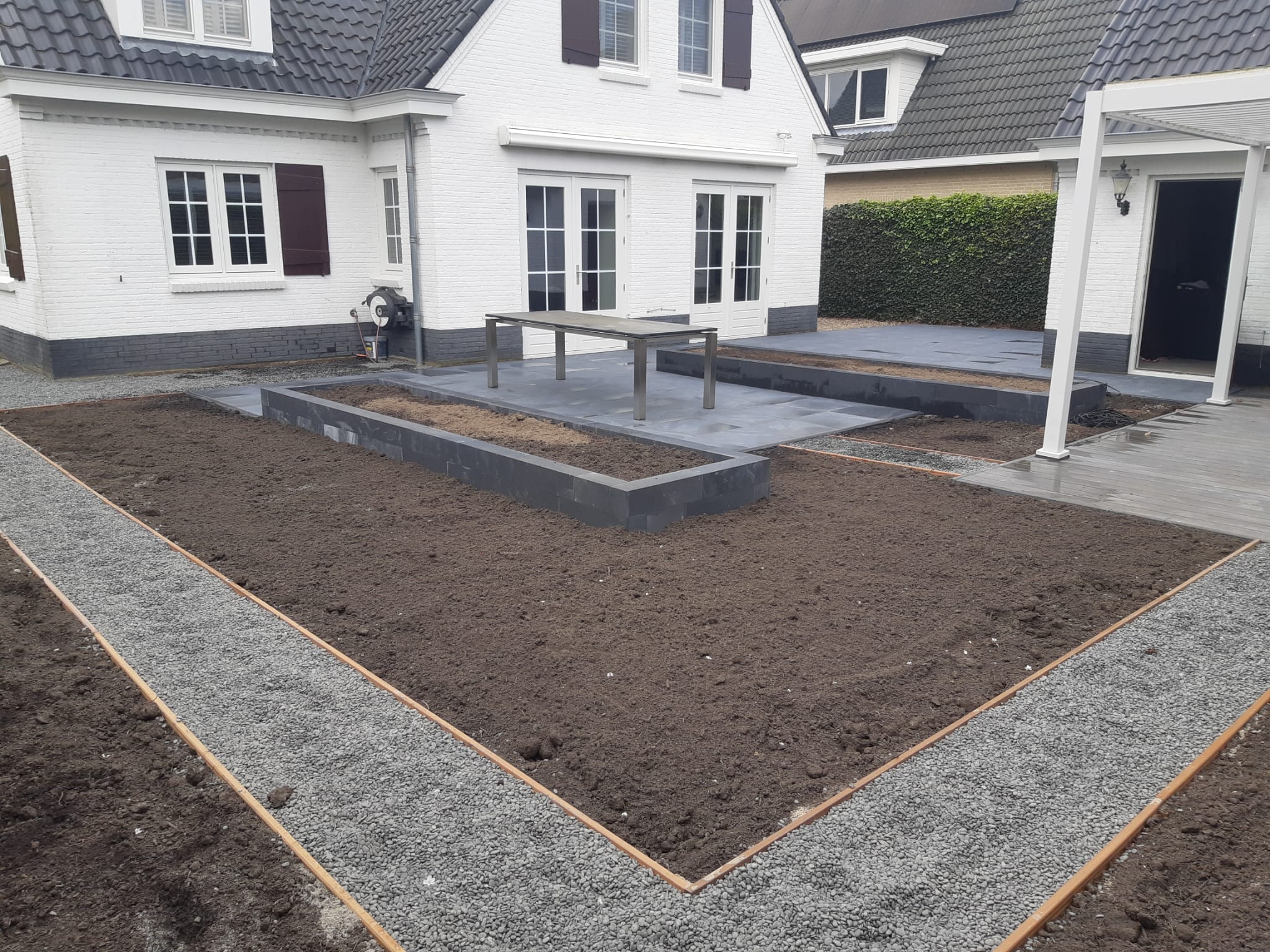 Tuin in aanleg bij een witte villa: grindpaden met stalen randen rond vers aangelegde borders en een terras